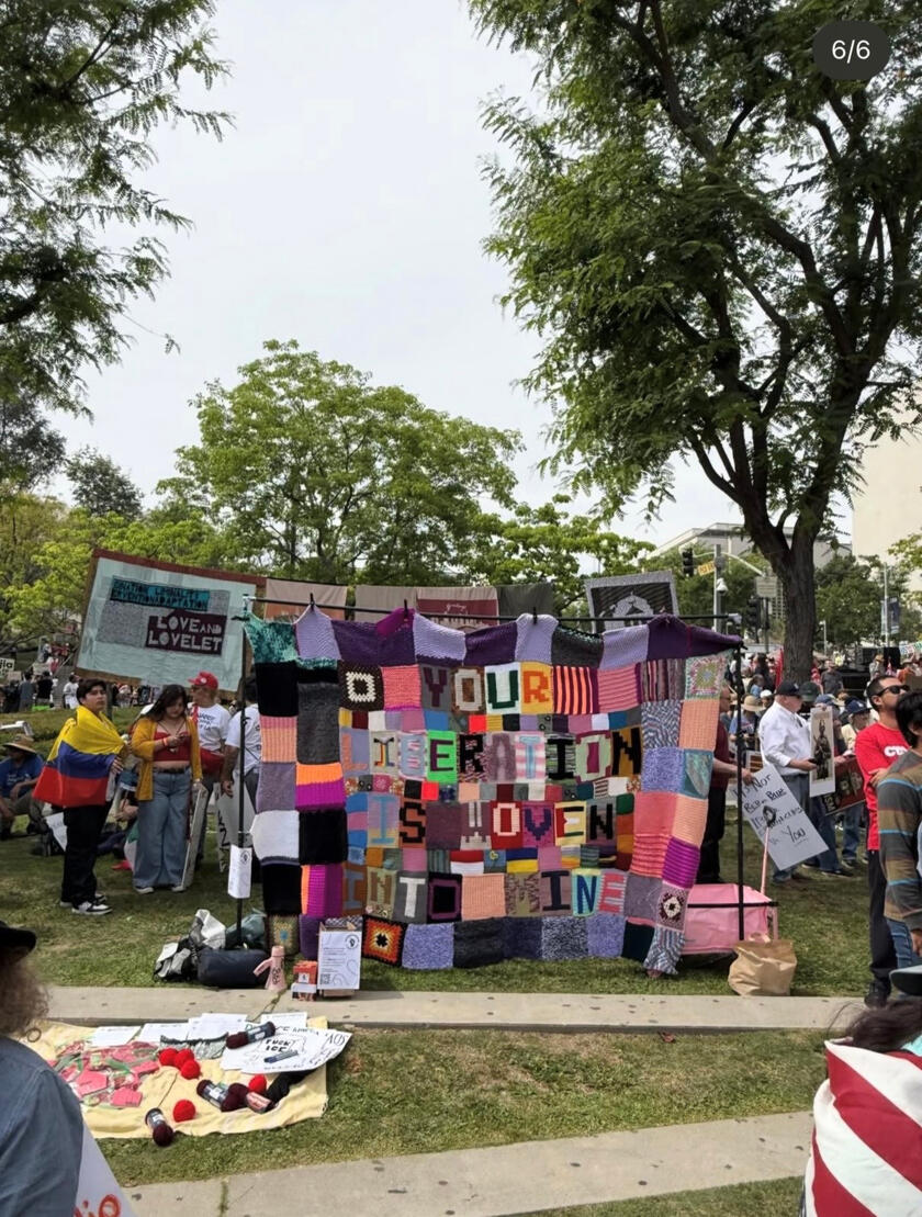 International collaborative tapestry, curated by and photographed by Raelee Sweet (@yarnivism), March 2026 Colorful knit and crochet tapestry quilt reading "Your Liberation is Woven Into Mine" in a green LA park